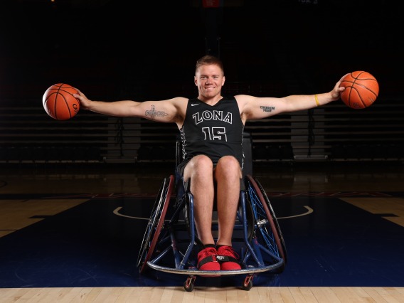 Hunter Pinke, an man using a wheelchair, holds two basketballs in both hands outstretched.  