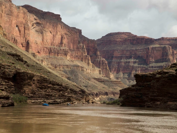 View of the Colorado river from the bottom of the Grand Canyon: two small rafts can be seen on the water in the distance and high cliffs rise in the background